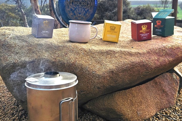Outdoor scene with a kettle steaming beside a rock with tea boxes and a mug on top.