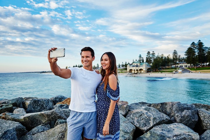 Couple taking a selfie on rocks by the ocean with a scenic beach and buildings in the background.