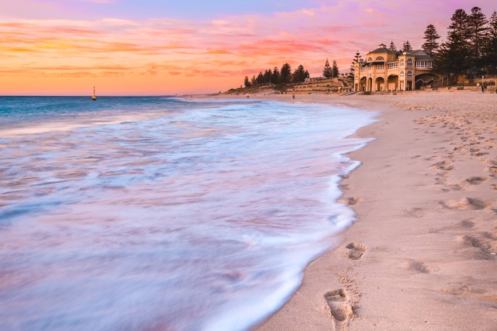 Sunset over beach with footprints and waves, a building onshore, and pastel sky.