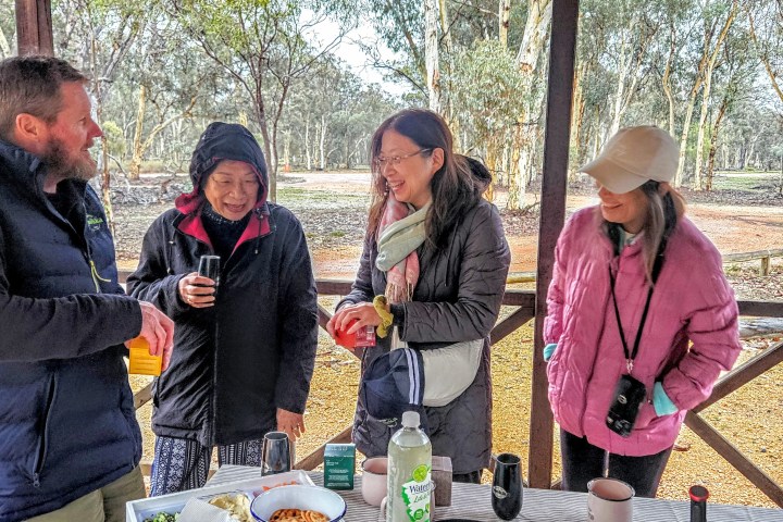 Four people chatting around a picnic table in a wooden shelter, with trees in the background.