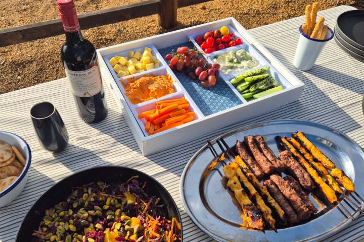 Outdoor table with wine, salad, grilled skewers, and veggie platter in a sunny setting.
