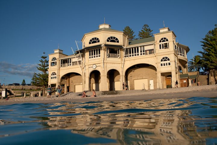 Historic beachfront building with arches, viewed from water, with people on sandy beach.