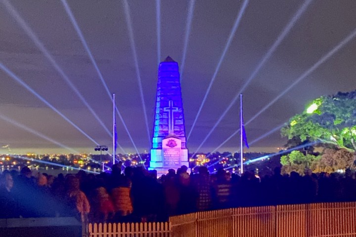 Illuminated obelisk with cross and spotlight beams, crowd gathered in foreground at night.