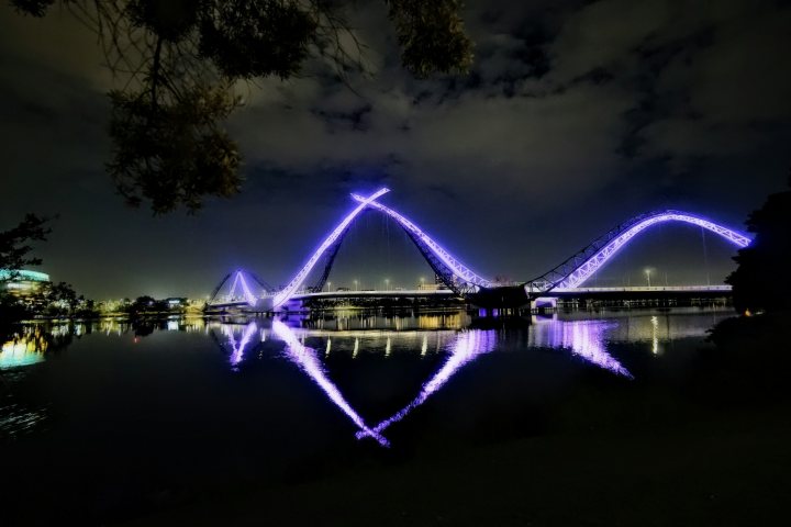 Illuminated bridge with purple lights reflected on a calm river at night.