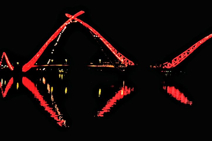 Illuminated red bridge with reflection on water against a dark background.