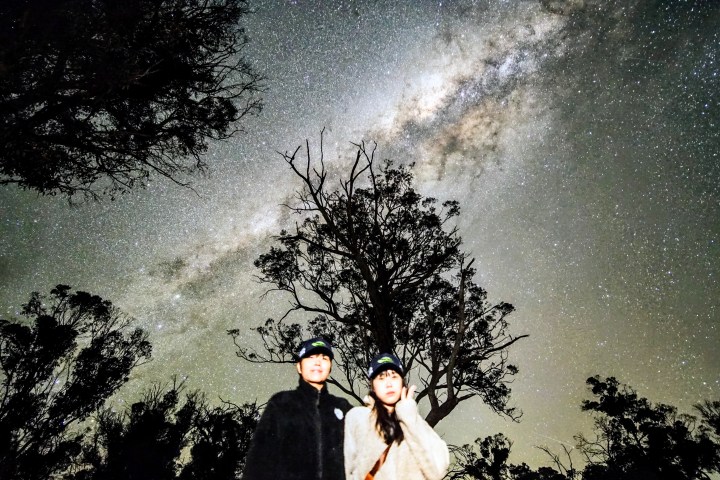 Two people under a night sky with the Milky Way visible, surrounded by silhouetted trees.
