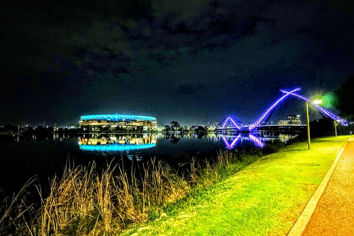 Night scene of stadium and bridge with blue lights reflected in water, pathway on right.