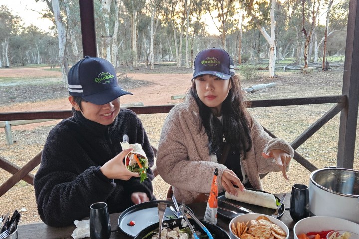 Two people in hats eating outdoors at a picnic table with various dishes.