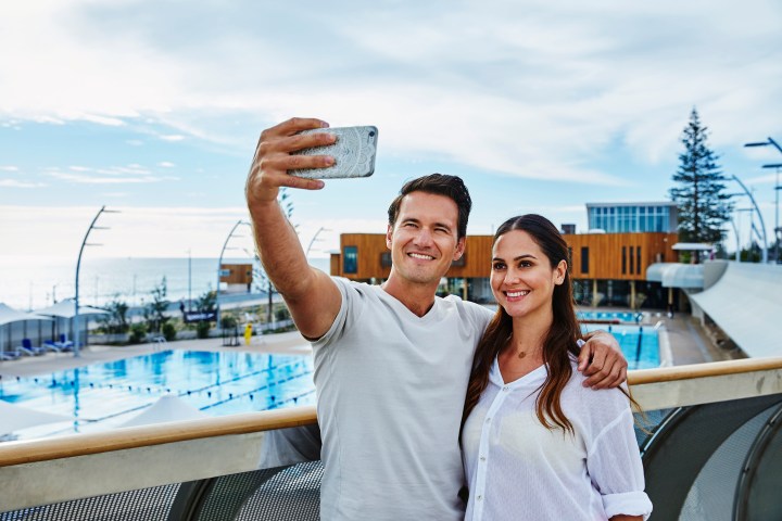 Couple taking a selfie in front of a pool and modern building by the sea.