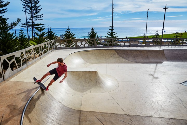 Skateboarder in red shirt riding in an empty skate park by the sea with clear sky.