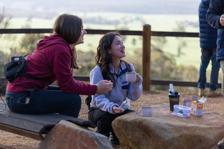 Two people outdoors, one sitting on a bench, the other kneeling, smiling, with mugs and snacks on a rock table.
