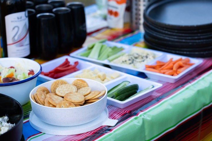 Table with snacks including crackers, cheese, cucumbers, vegetables, and wine on a colorful tablecloth.