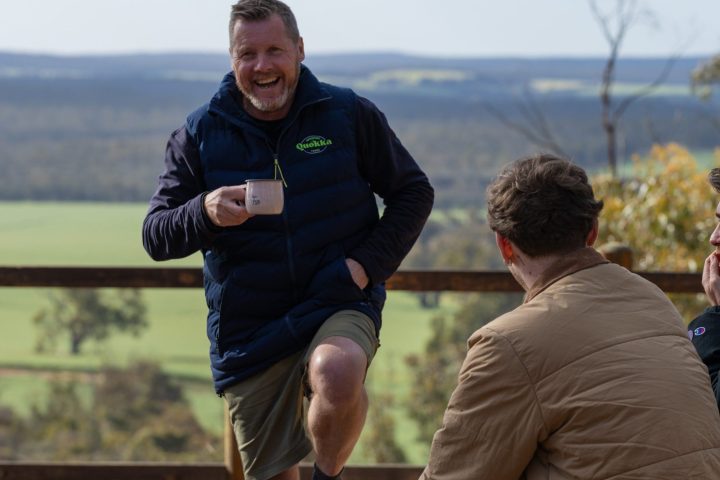 Three people outdoors; one standing, laughing, holding a mug. Two seated on rocks, all in casual clothing.