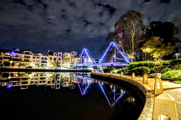 Night view of a brightly lit bridge reflecting in a calm river beside a paved walkway.