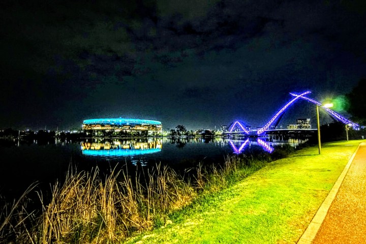 Lit stadium and bridge reflected on water at night, with a lit pathway on the right.