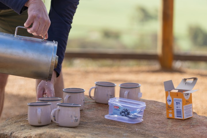 Person pouring beverage into metal mugs on a rock with sugar box and container.