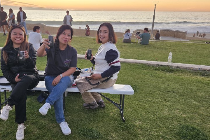 Three people sitting on a bench, holding drinks, with a beach sunset view in the background.