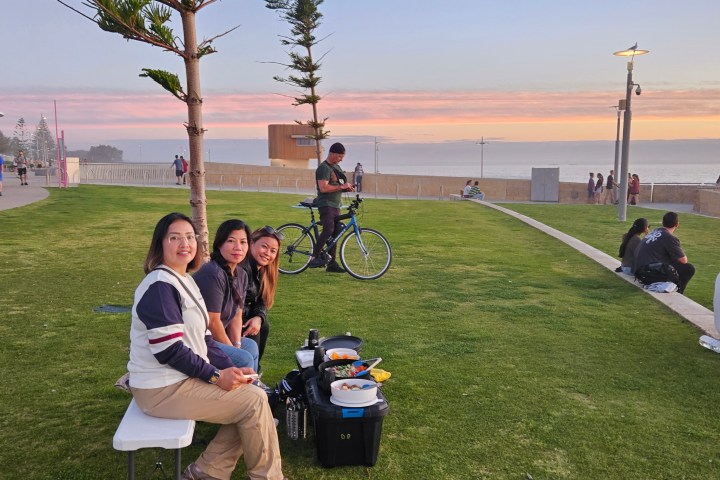 Three people seated on a lawn near a path, with a sunset and ocean in the background, others walking nearby.