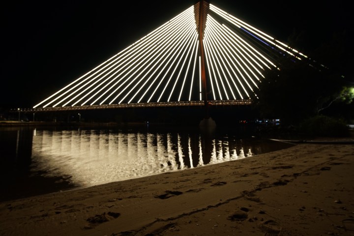 Illuminated bridge at night with reflections on water and sandy shore in foreground.