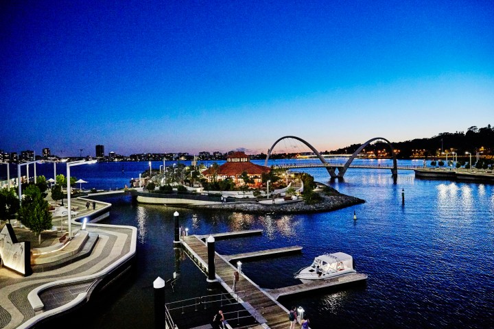 Harbour scene at dusk with curved bridge, boats, and city lights.
