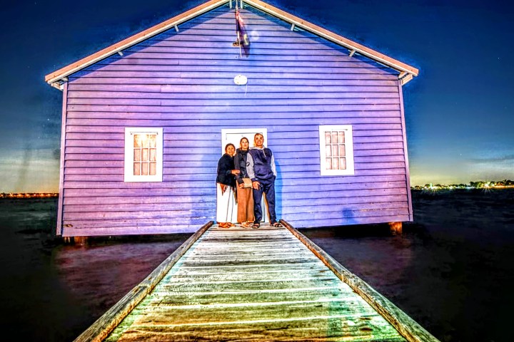 Three people stand on a pier in front of a blue house over water at night.