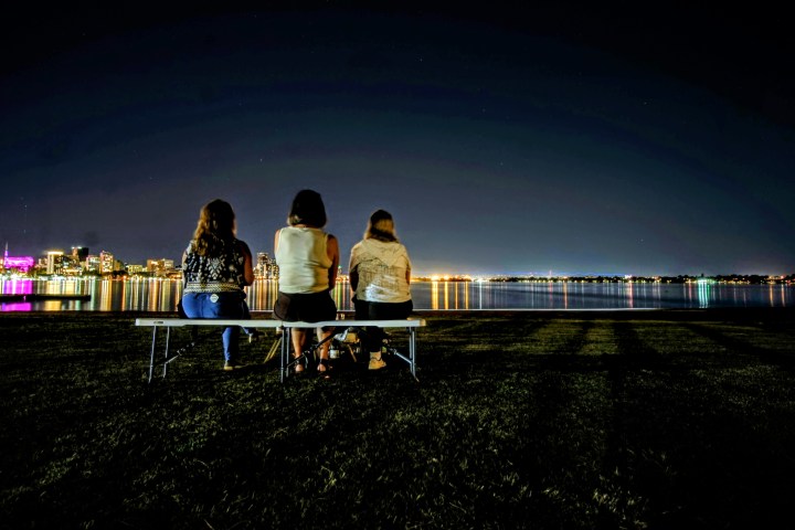 Three people sitting on a bench by a river at night, with city lights reflecting on the water.