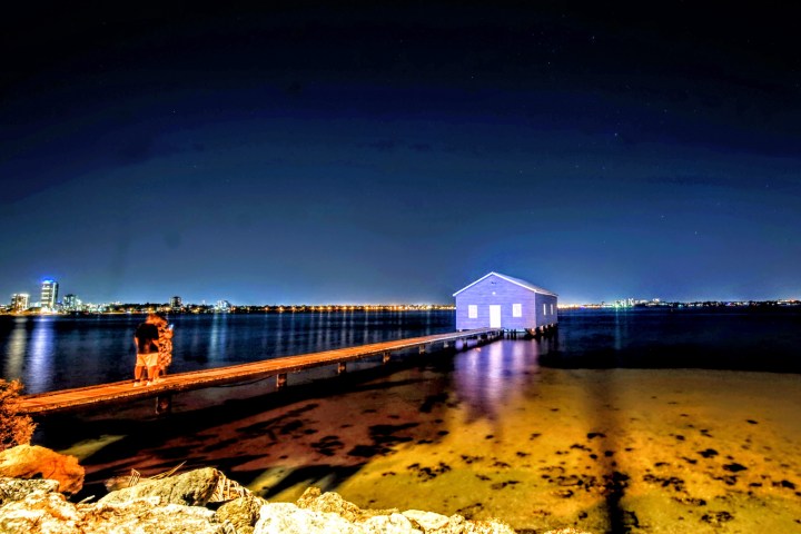 Blue boathouse on water with pier at night, city lights in background.